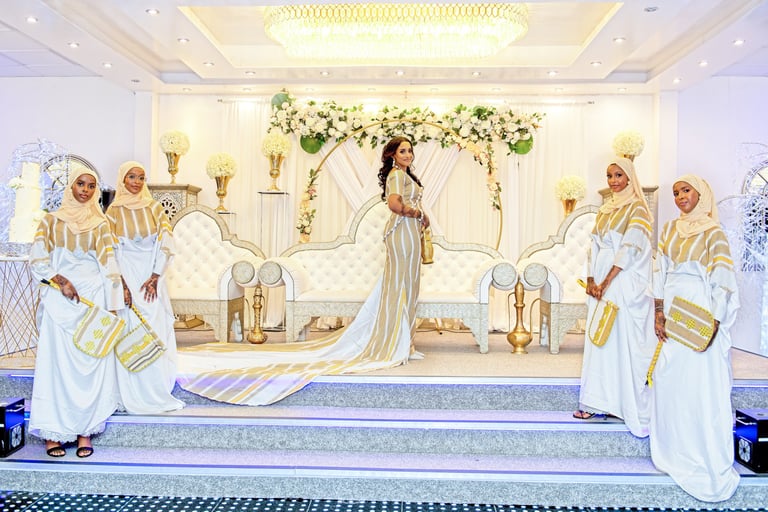 Somalian bride in centre, bridesmaids are next to her all in traditional white & gold outfits looking at camera