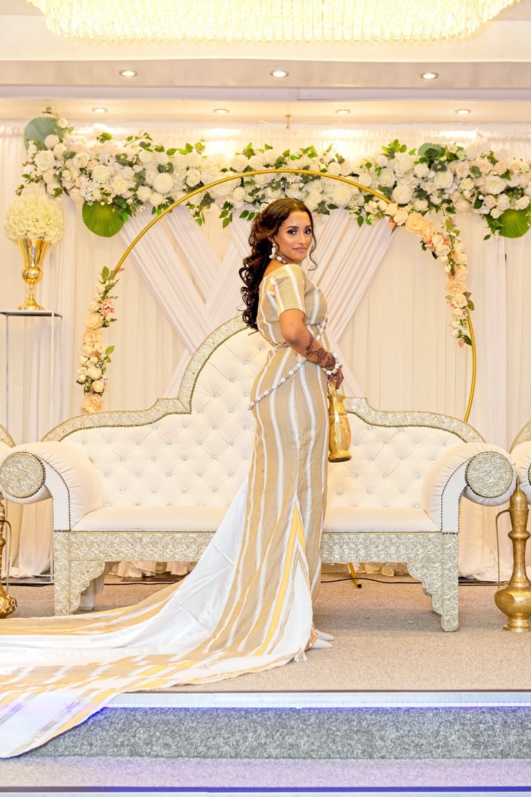 Somalian bride dressed in traditional white & gold outfit on white floral background, looking at camera