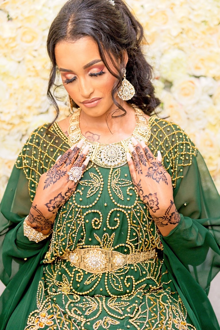 Somalian bride in traditional green dress with gol beads putting gold necklace on, blurry floral background