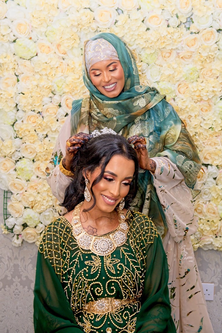 Somalian mother of bride helping ride get ready, both in traditional green & gold outfits, blurry floral background 