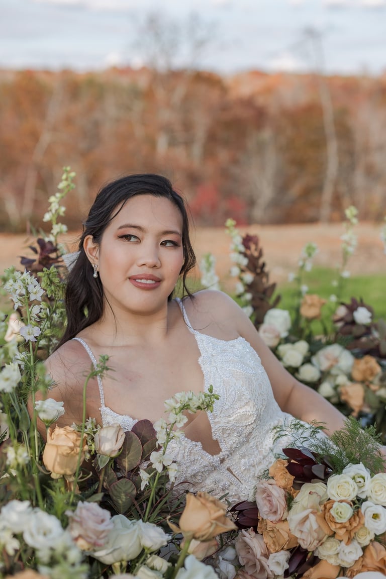 Asian bride with romantic makeup and long dark hair in half up hairstyle, wearing lace gown and surrounded by roses outdoors.