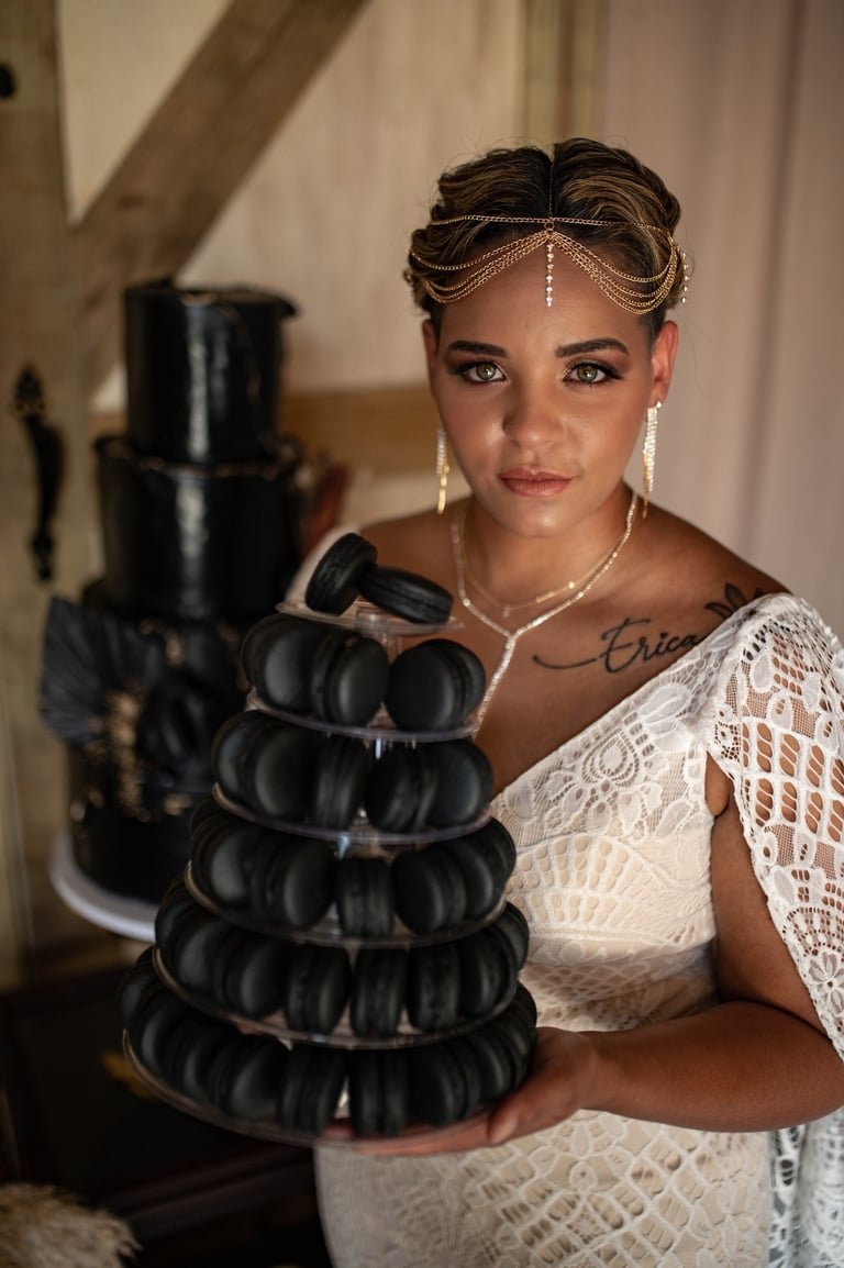 Bride with natural glam makeup and braided updo wearing lace gown and gold headpiece, holding black macaron tower at wedding.