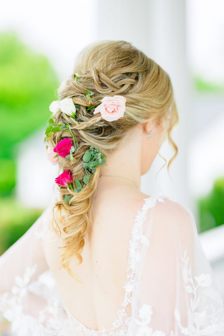 Bride with blonde braided hairstyle decorated with pink roses and greenery, romantic floral bridal look from back view.