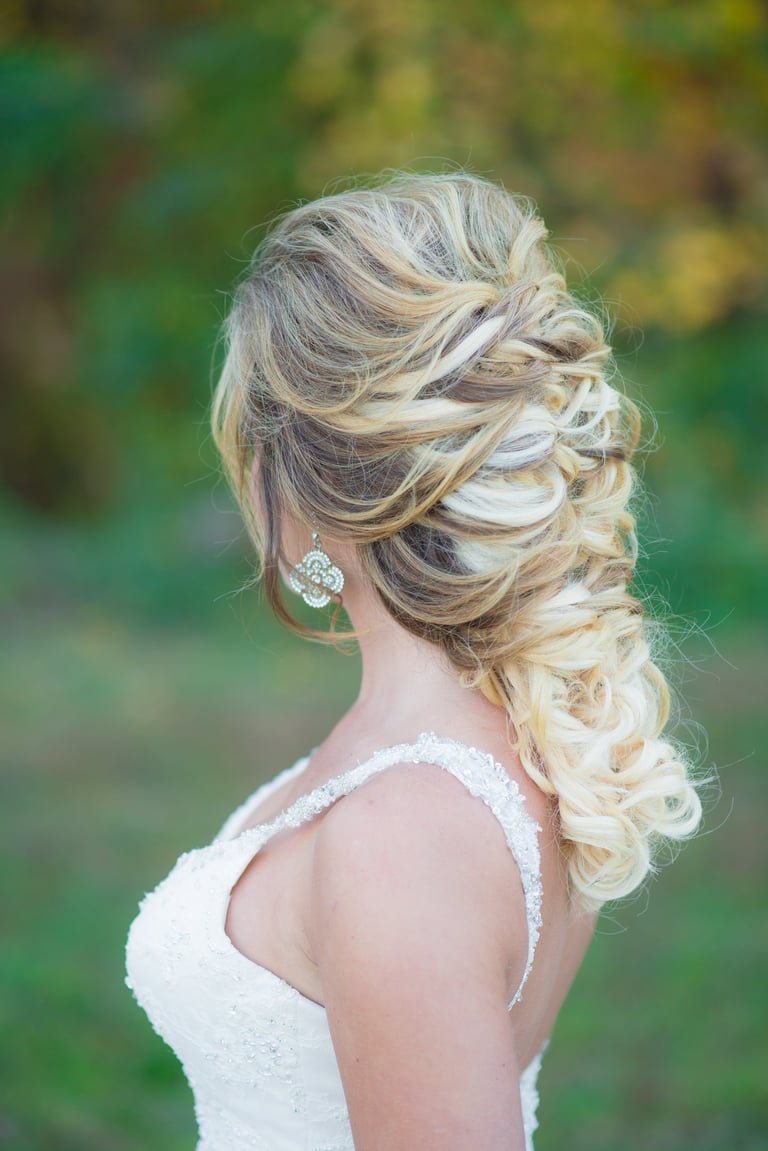 Bride with voluminous blonde braided wedding hairstyle wearing jeweled earrings and lace gown, elegant outdoor bridal look.