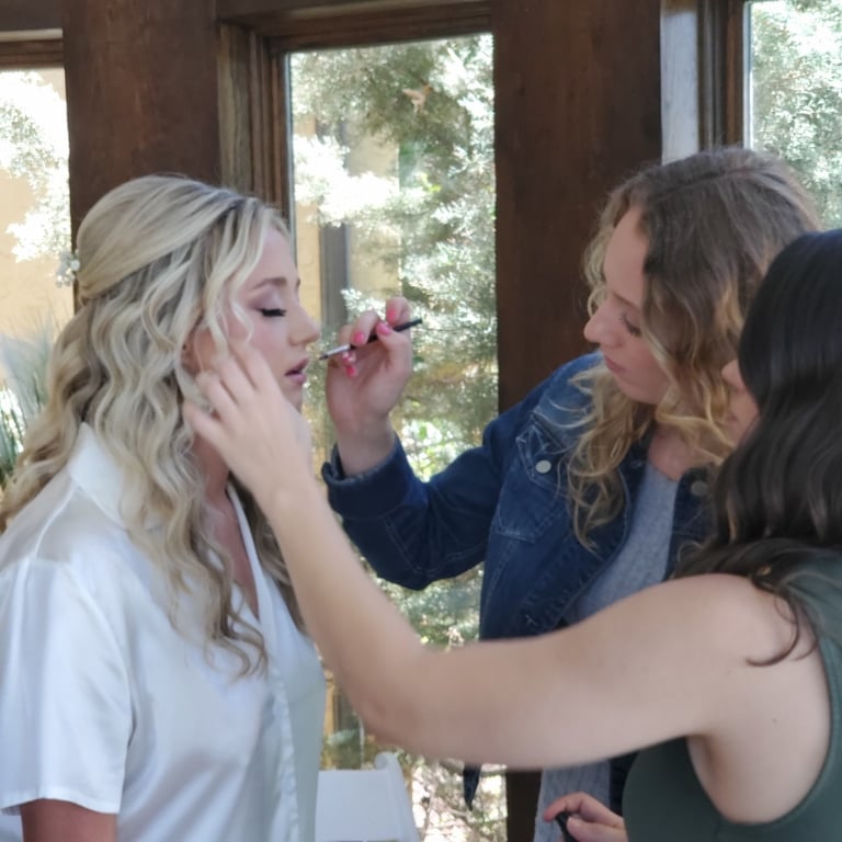 Bride with long blonde waves in a silk robe receives final touch ups from stylists, highlighting her wedding hair and makeup.