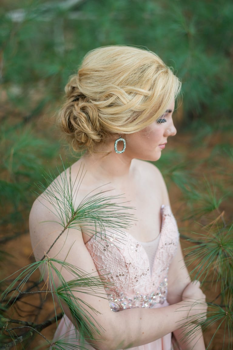 Bride with blonde textured updo hairstyle wearing sequin pink dress and earrings, standing among pine trees outdoors.