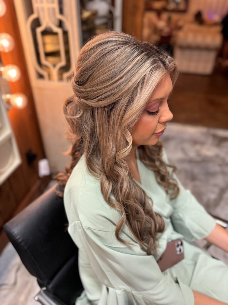 Bride with blonde hair in half up half down soft waves, wearing sage robe and glowing makeup while preparing for wedding day.