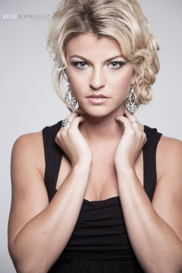 Woman with blonde textured side updo hairstyle and dramatic eye makeup wearing black dress and statement chandelier earrings.