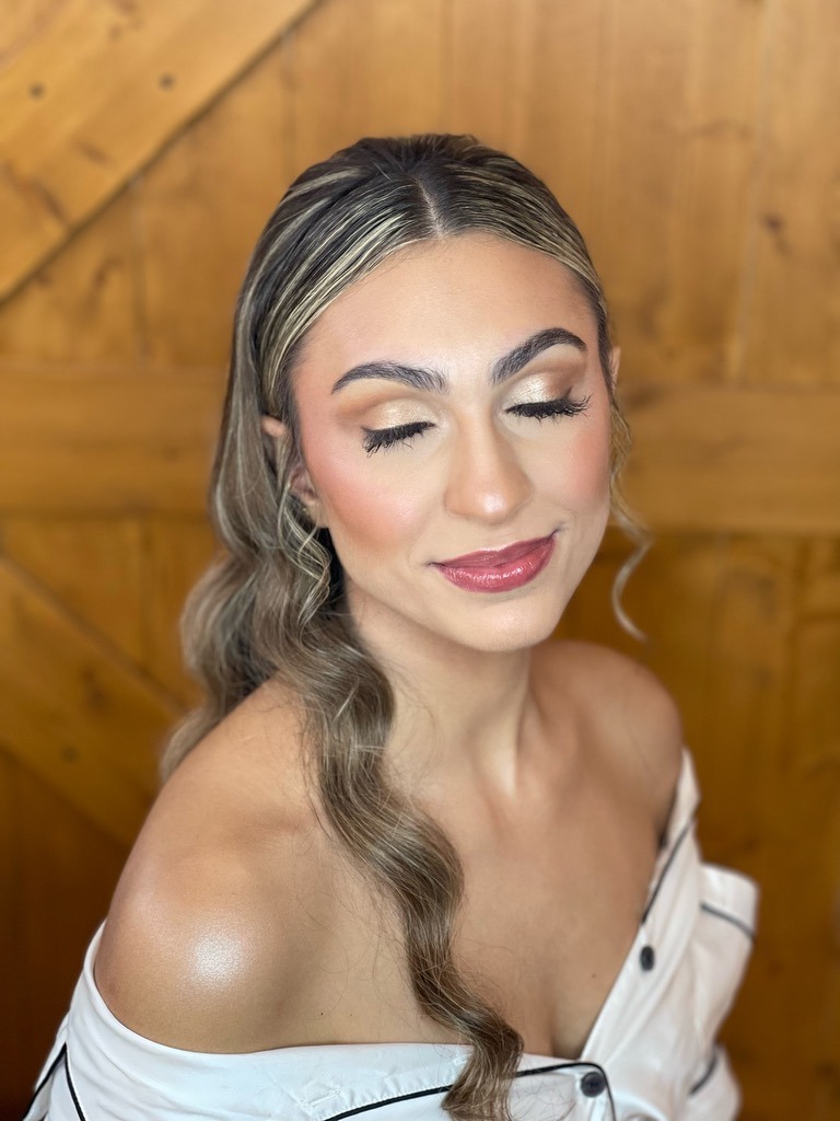 Bride with glam waves hairstyle, glowing skin, neutral eyeshadow, long lashes, and glossy lips wearing an off-shoulder gown.