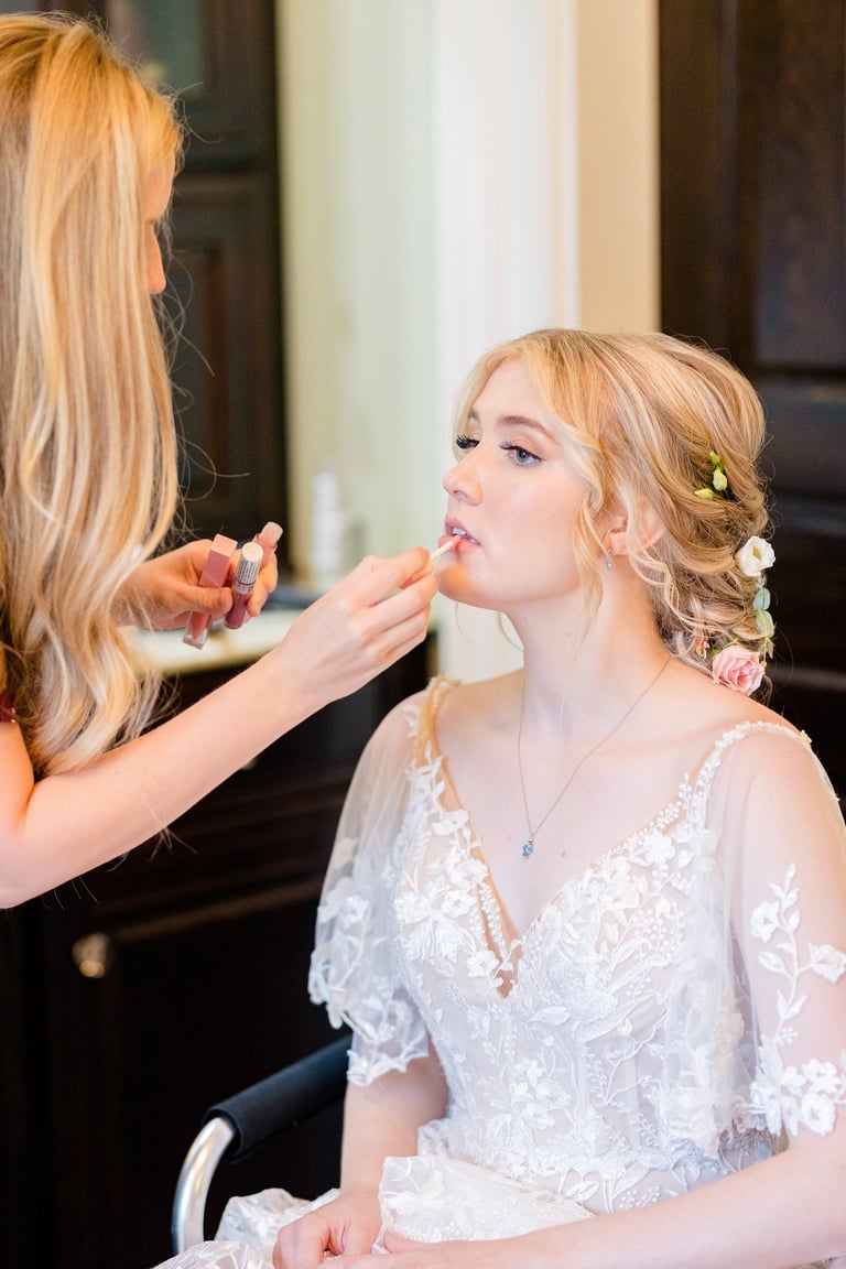 Bride in lace gown with floral updo receives lip gloss from makeup artist, highlighting wedding hair and bridal makeup.