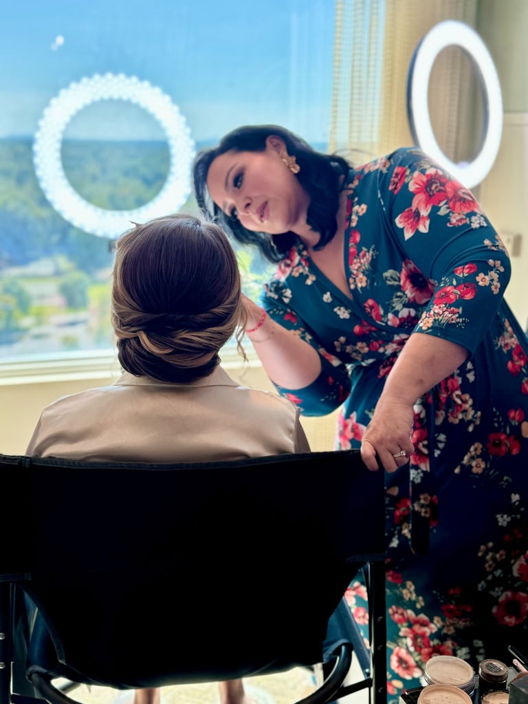 Makeup artist refines the bride's makeup beside a window; twisted low updo and ring light showcase elegant wedding hair.