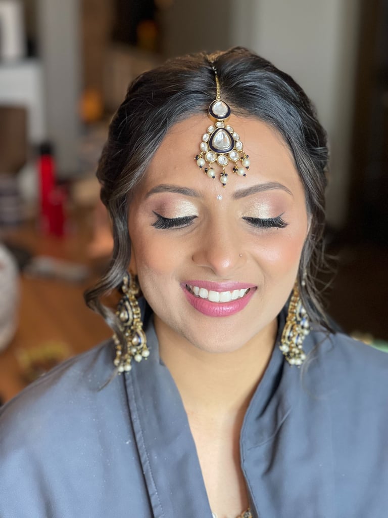 Indian bride with eyes closed, soft-glam wedding makeup, pearl maang tikka, pink lips, long lashes, and ornate earrings.