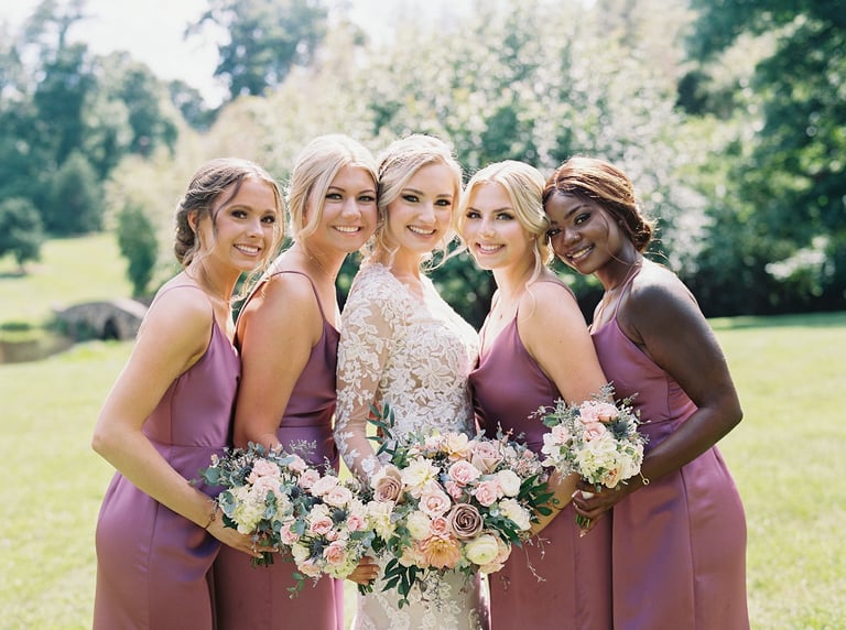 Bride with soft updo and natural wedding makeup smiles with bridesmaids in mauve dresses holding pastel flower bouquets.