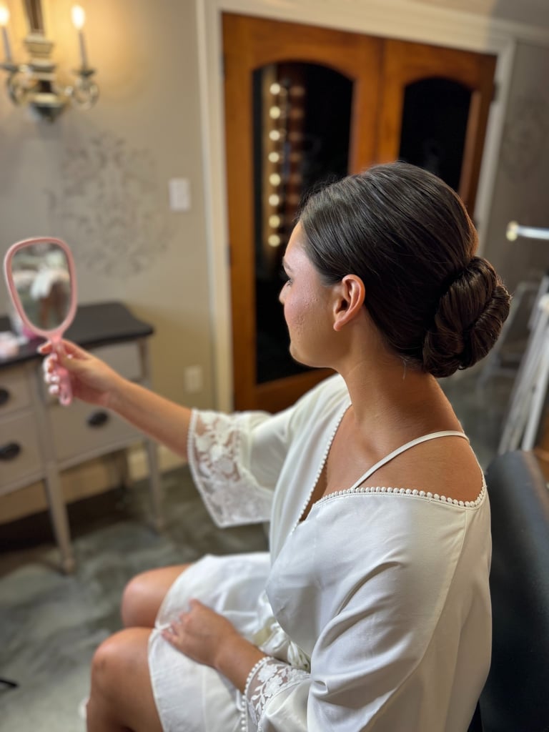 Bride in white robe holding mirror admiring sleek low bun hairstyle, elegant polished look while getting ready for wedding.
