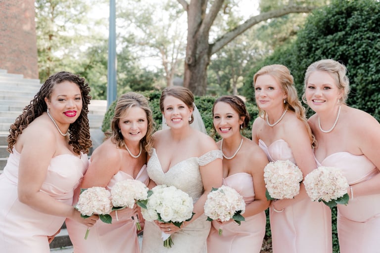 Bride with white bouquet smiling with bridesmaids in blush dresses, wearing elegant down hairstyles and holding hydrangeas.