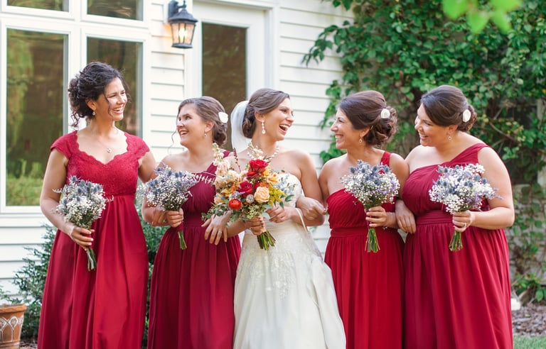 Bride with colorful bouquet smiling with bridesmaids in red dresses, each wearing elegant updo hairstyles, holding bouquets.