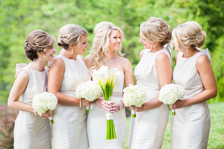 Bride with long blonde hair and bridesmaids in matching white dresses, all with elegant updos, holding white bouquets.