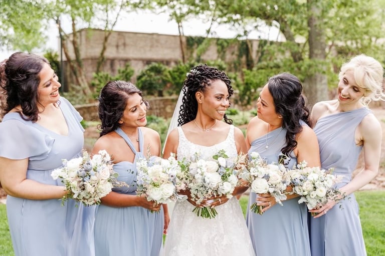 Bride in lace gown with bridesmaids wearing light blue dresses, holding white and pastel bouquets at an outdoor wedding.