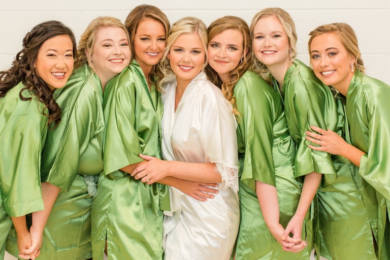 Bride in white satin robe surrounded by bridesmaids in matching green robes, all getting ready together on wedding morning.