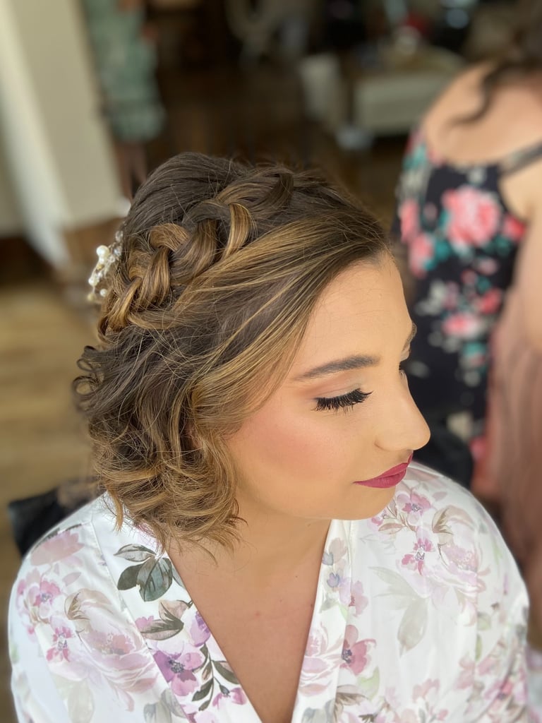 Bride with short hair styled in braided crown and curls, wearing floral robe and elegant makeup for wedding day preparation.