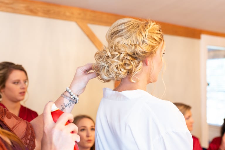 Stylist applies hairspray to bride’s braided curly updo, capturing an elegant wedding hairstyle preparation.