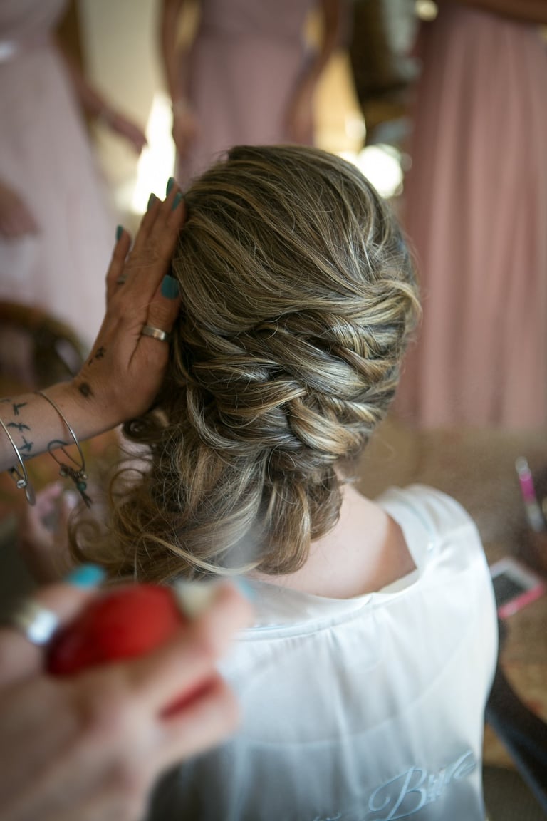Close-up of bride’s braided updo as stylist applies hairspray, highlighting elegant wedding updo with soft curls and detail.