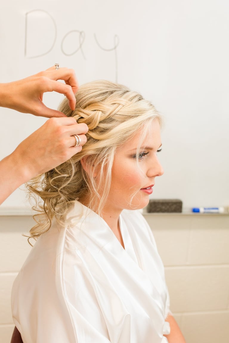 Bride with blonde hair styled into a braided updo receives final touches, showcasing soft curls and elegant bridal makeup.