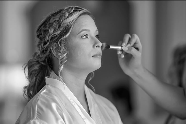 Bride with braided updo and silk robe receives bridal makeup touch in a candid black and white getting ready moment.