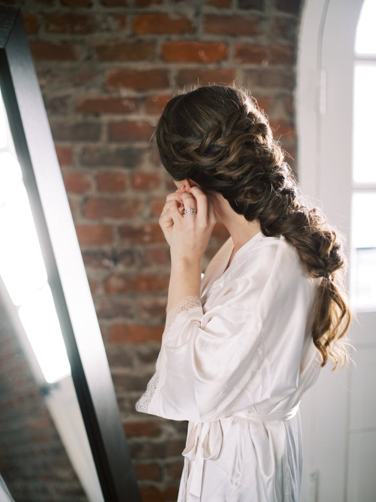 Bride in silk robe adjusting earring with long braided wedding hairstyle, romantic look while getting ready by brick wall.