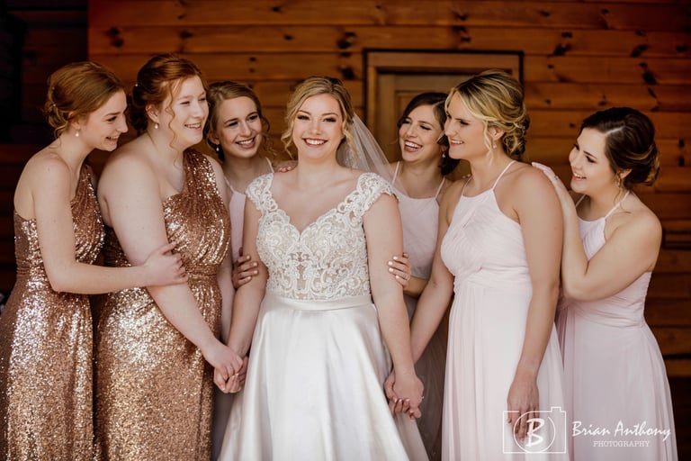 Bride in lace gown with elegant updo and soft wedding makeup, surrounded by bridesmaids in blush and sequin dresses.