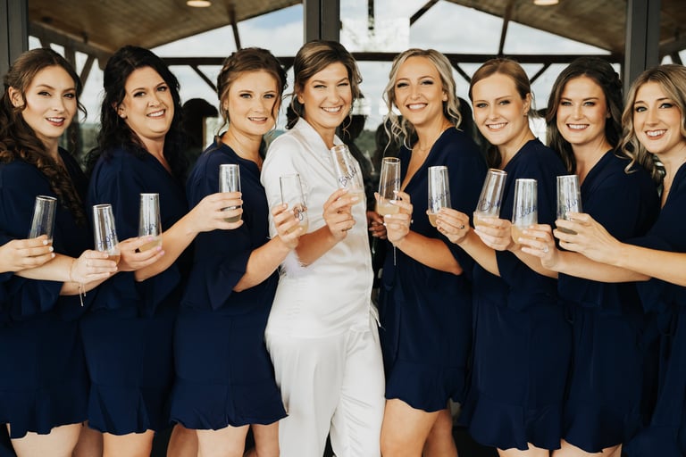 Bride in white pjs with bridesmaids in navy robes holding champagne glasses, showing elegant wedding hair and natural makeup.