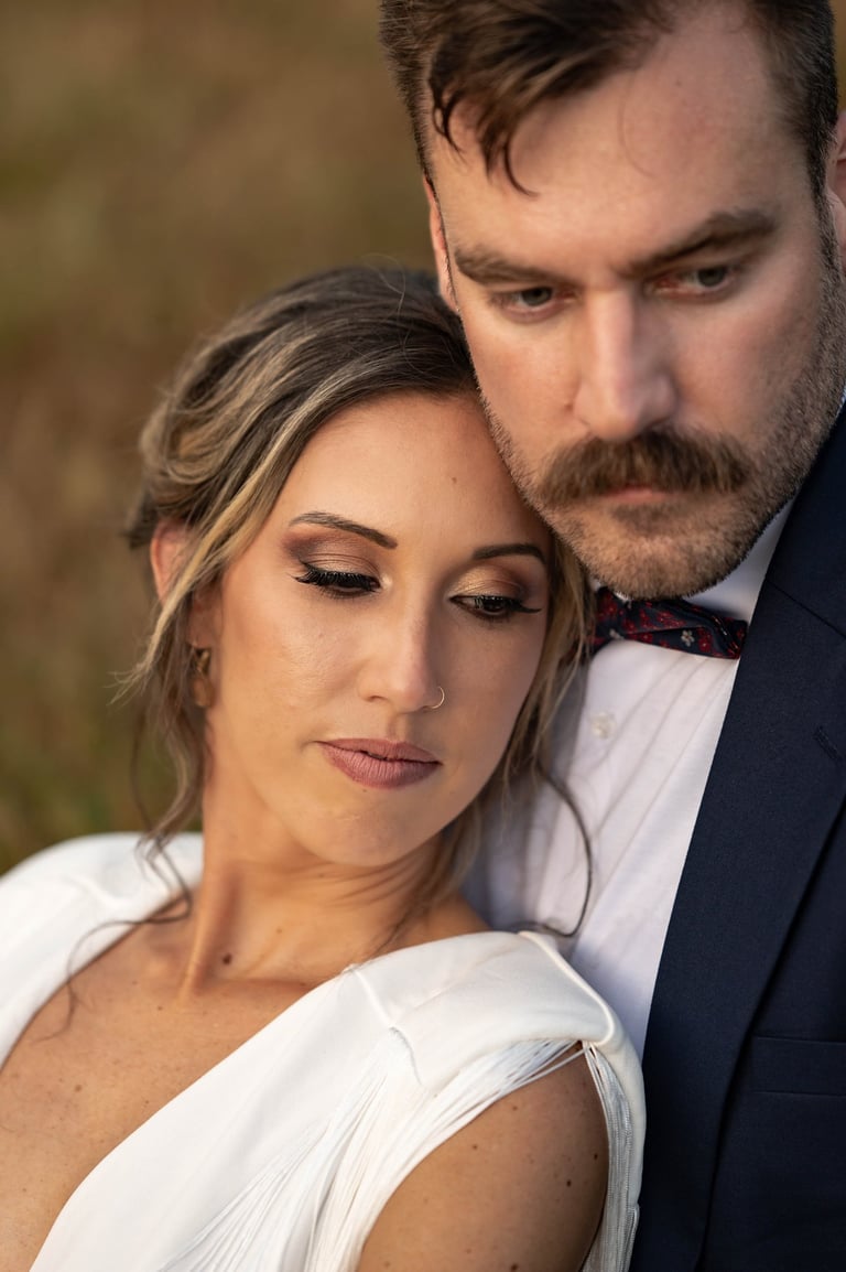 Bride with dramatic wedding makeup featuring bold eyeshadow and nude lips, leaning against groom in navy suit for portrait.