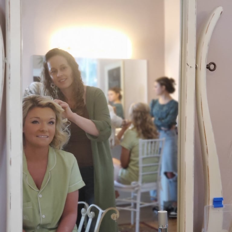Bride in green pajamas smiles in mirror while stylist curls her hair, capturing a candid wedding getting ready moment.