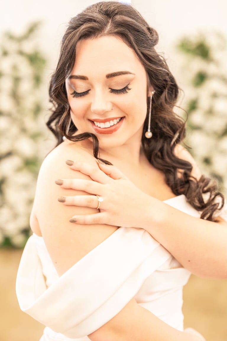 Bride with glamorous wedding makeup, long curled hair, pearl drop earrings and off shoulder gown, smiling with eyes closed.