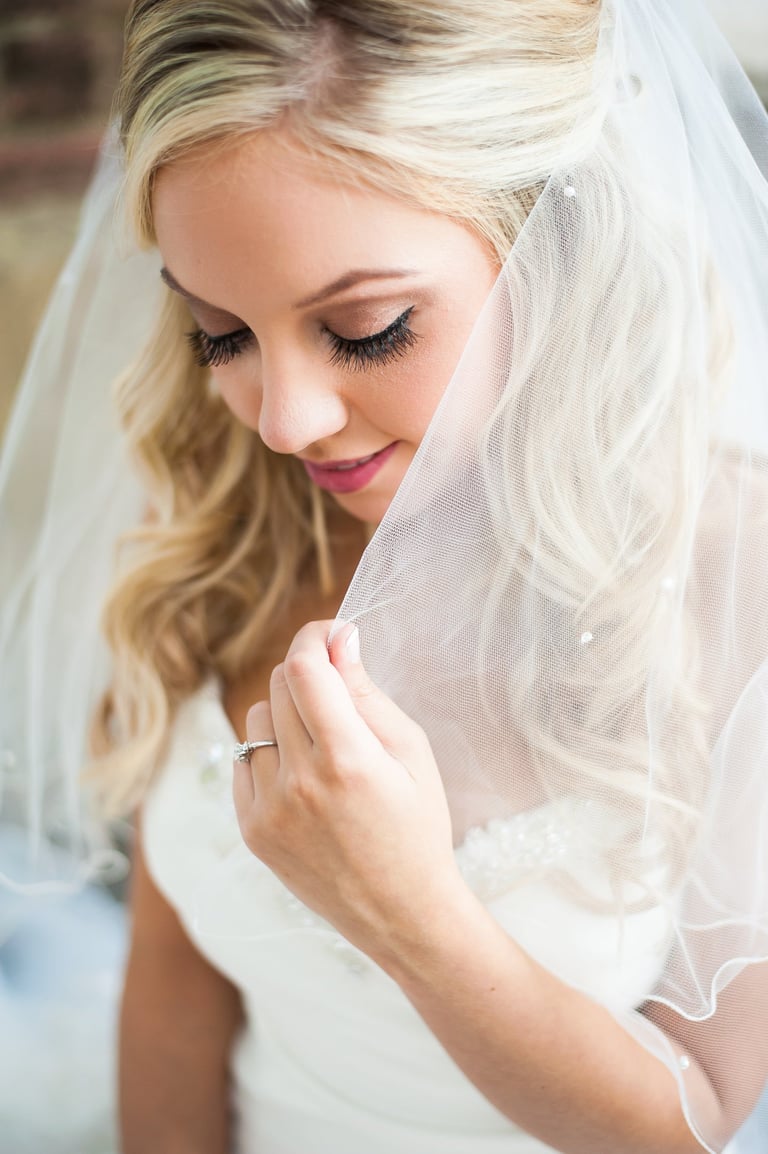 Bride with glamorous eye makeup and long lashes holding veil, wearing lace gown with curls, elegant bridal close-up portrait.
