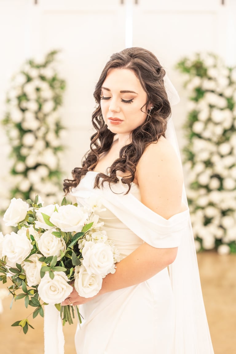 Bride with dark hair styled in half up half down curls wearing off shoulder gown, holding white rose bouquet at wedding.