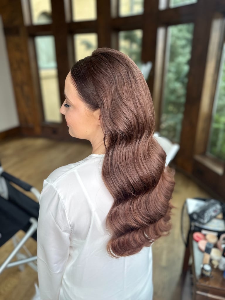 Bride with long brown hair styled in classic Hollywood glam waves, wearing white robe while getting ready on wedding morning.