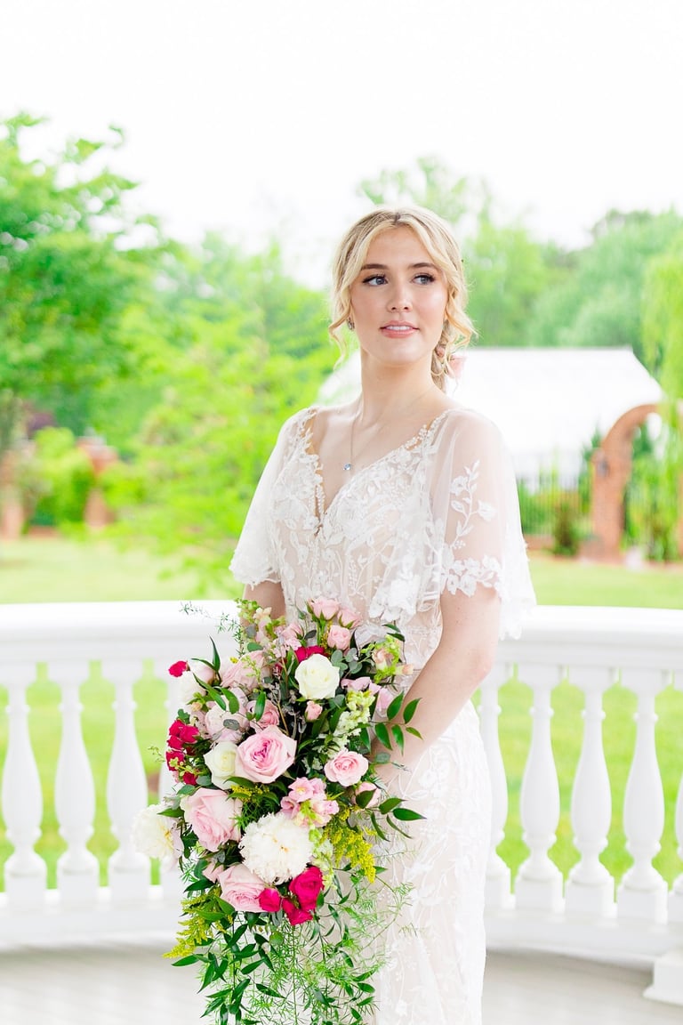 Bride with natural makeup and soft updo hairstyle holding a cascading bouquet of pink and white flowers on a porch with green
