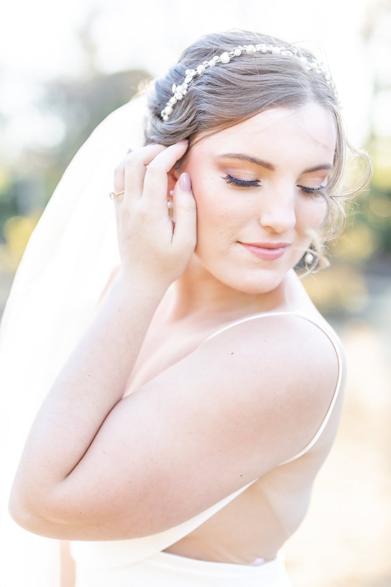 Bride with natural makeup and soft updo hairstyle wearing pearl headband and veil, captured in elegant outdoor portrait.