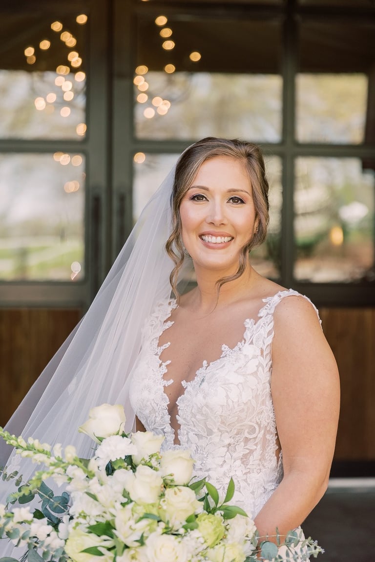 Bride with natural glowing makeup and soft updo hairstyle wearing lace gown, holding white floral bouquet with veil draped.