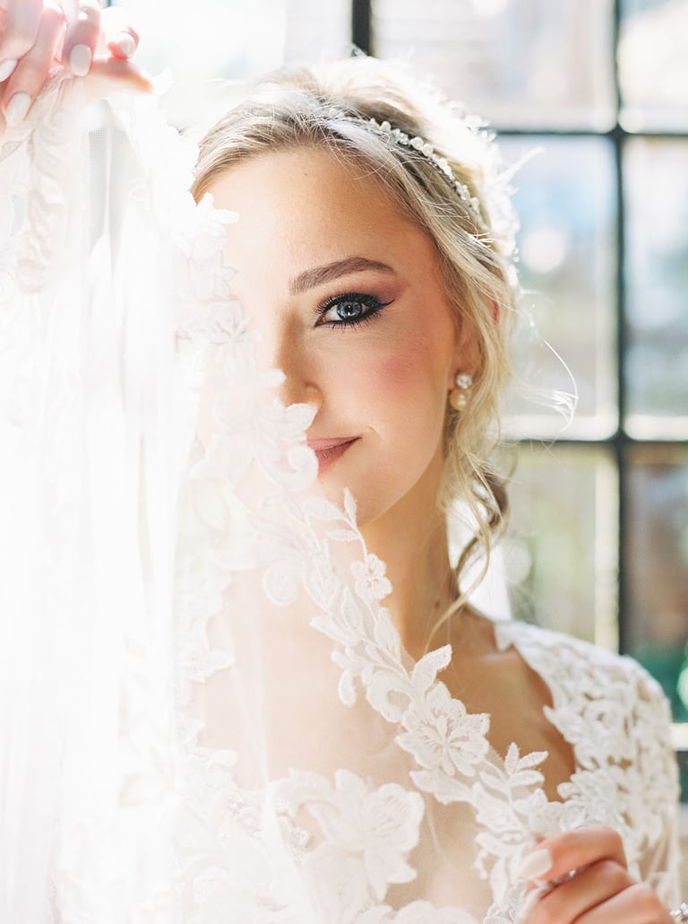 Bride with romantic wedding makeup holding lace veil in soft natural light, showing elegant eye makeup and flawless skin.