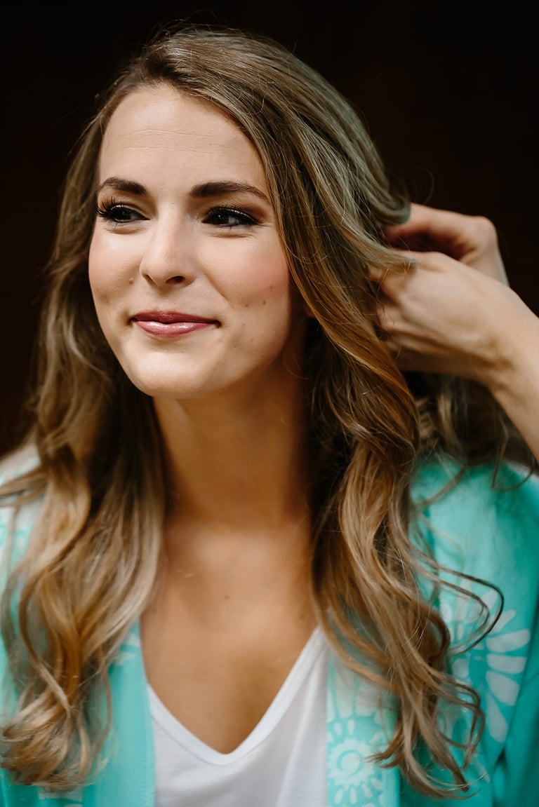 Bride in a teal robe smiles as a stylist shapes soft waves, showcasing natural bridal makeup and romantic wedding hair.