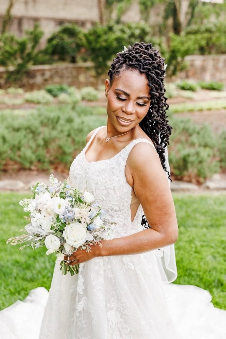Bride with long twisted locs hairstyle wearing lace gown and holding white floral bouquet, smiling outdoors on wedding day.