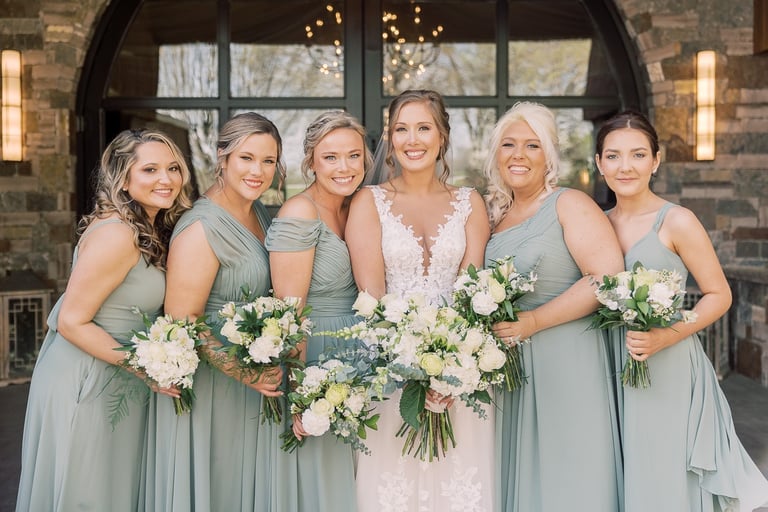 Bride with five bridesmaids in sage green dresses holding white bouquets, showcasing natural bridesmaid makeup looks.