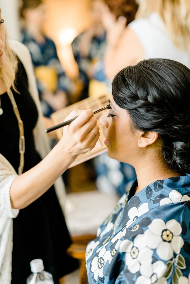 Bride with braided updo in a floral robe receives eyeshadow from makeup artist, a candid wedding hair and makeup prep moment.