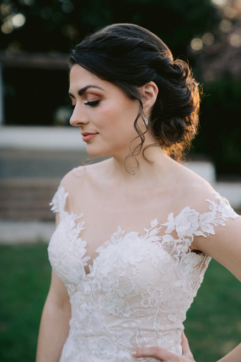 Bride with dark hair styled in elegant twisted updo wearing lace wedding dress, natural makeup and soft curls framing face.