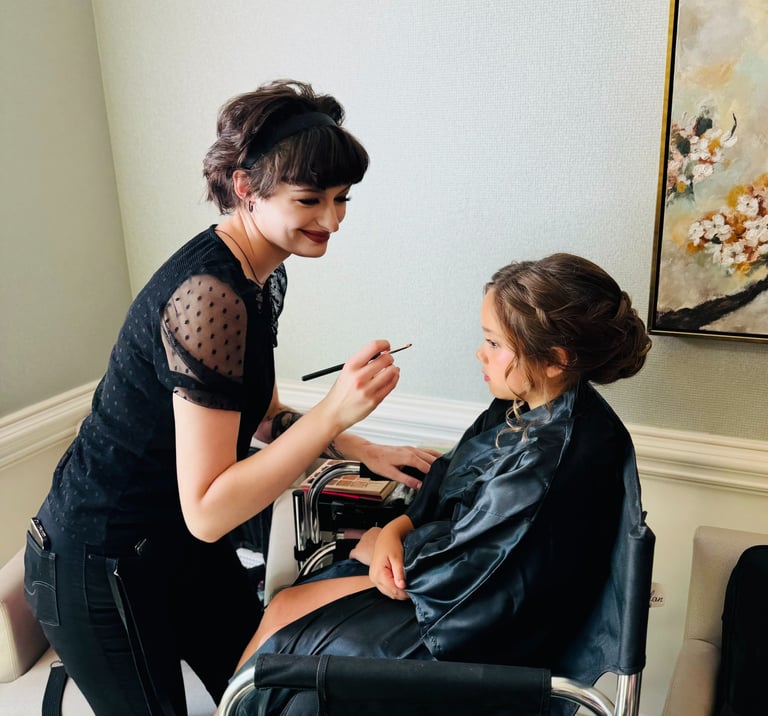 Makeup artist adds blush to a flower girl with a braided updo, capturing sweet wedding hair and makeup getting ready moment.