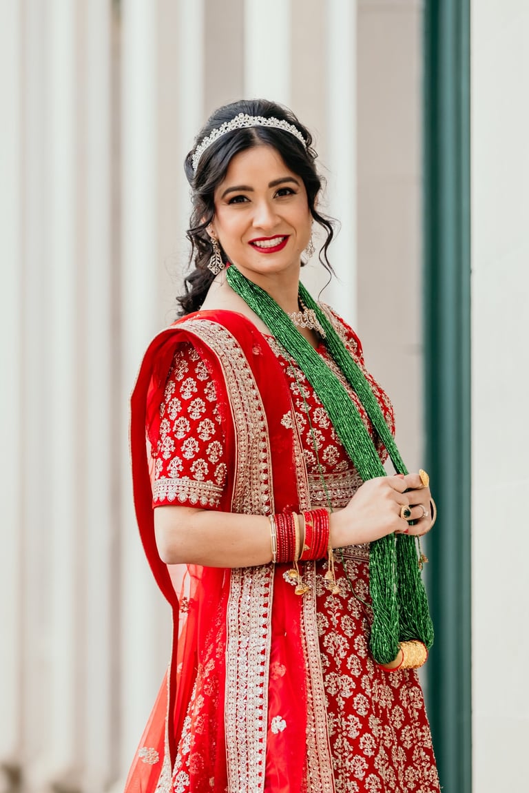 Indian bride with bridal makeup wearing red lehenga, green necklace, tiara, and bold red lipstick for traditional wedding.