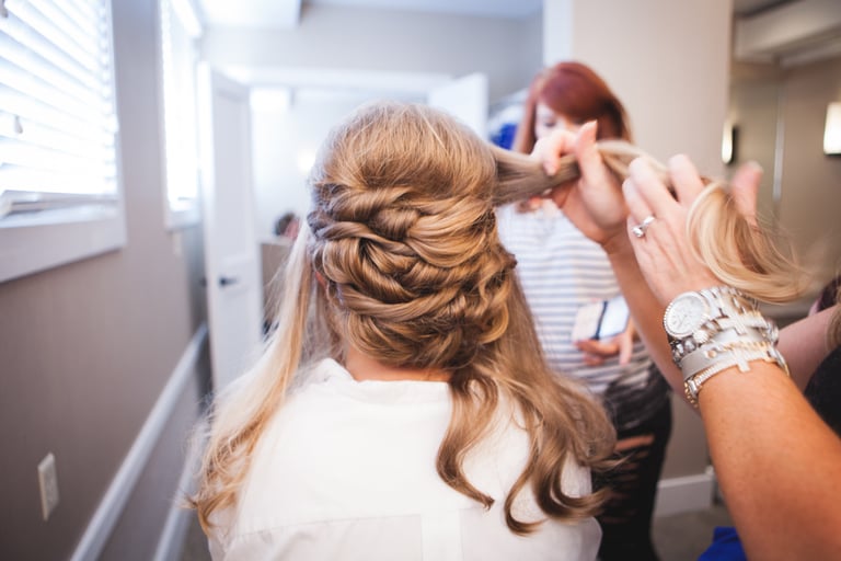 Stylist creates an intricate half up braided wedding hairstyle, weaving curls into a polished look as the bride gets ready.