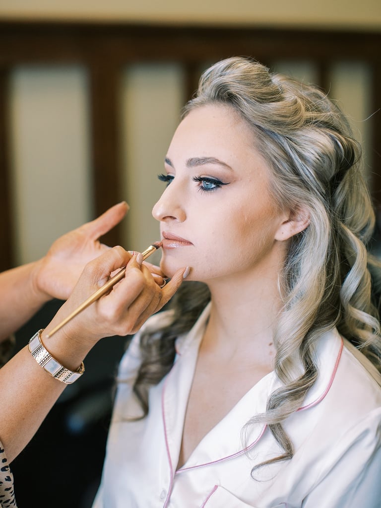 Makeup artist applies lipstick to the bride with soft waves and winged liner, highlighting polished wedding hair and makeup.